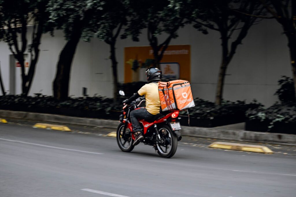 A delivery man riding a motorcycle with a thermal bag on a city street during the day.