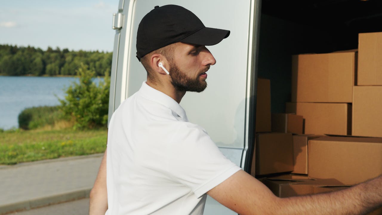 Courier wearing earphones loading packages into a van by a lakeside road.