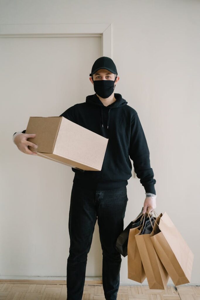 pexels-photo-6612497 A delivery man wearing a mask holds a box and shopping bags indoors.